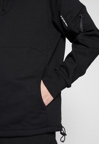 Černá mikina s texturovanou látkou, bočními kapsami, límcem na čtvrtinový zip a kapsou na zip na rukávu se šňůrkou.