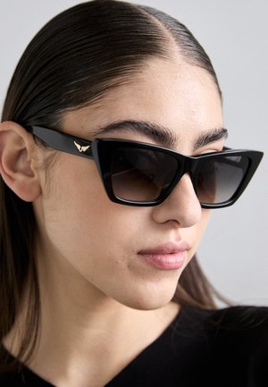 Óculos de sol estilo gato preto com lentes escuras, apresentando um detalhe dourado na lateral. A armação tem um acabamento mate e design angular.