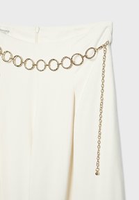 Jupe blanche avec une ceinture décorative en chaîne dorée composée de maillons circulaires texturés et une extrémité de chaîne réglable qui pend.
