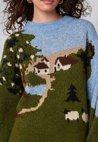 Pletený svetr s krajinovým designem v zelených a modrých odstínech, zobrazující stromy, domy, řeku a ovce. Měkká textura, kulatý límec.
