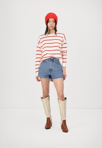 Pulover cu dungi roșii și albe, șorturi din denim albastru și boots de cowboy bej cu accente crem. Modelul poartă o căciulă tricotată roșie aprinsă.