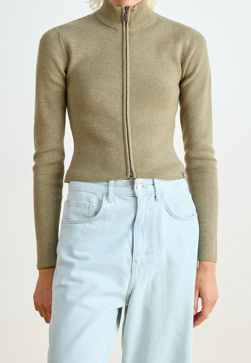 Personne portant un pull zippé côtelé vert olive ajusté et un jean taille haute bleu clair sur un fond uni.