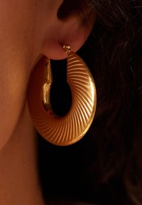 Gouden oorbel in de vorm van een ring met een geribbelde textuur en een cirkelvorm, voorzien van een veilige sluiting met een pen en die het licht reflecteert dankzij de gepolijste afwerking.