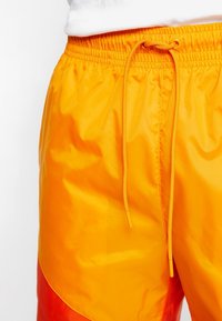 Žluté a oranžové nylonové šortky s možností stažení v pase. Látka má hladkou texturu a jednobarevný design.