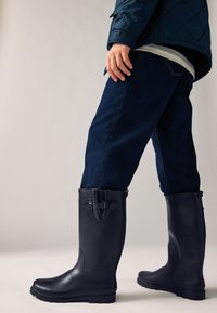 Schwarze kniehohe Gummistiefel mit matter Oberfläche, rechteckiger Zehenpartie, Schnallendetail am Schaft und dicker, strukturierter Sohle, kombiniert mit dunklen Jeans.