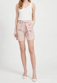 Woman wearing a white sleeveless top, high-waisted pink shorts with a tied belt, and white pointed-toe heels, standing against a plain background.