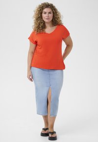 Camiseta naranja de cuello en V, de mangas cortas, combinada con una falda lápiz de mezclilla azul claro con una abertura frontal. Sandalias planas negras.