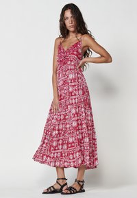 Rotes Maxikleid mit weißem, kunstvollem Muster, Spaghettiträgern, tailliertem Oberteil und gestuftem, rüschendem Saum. Kombiniert mit schwarzen Sandalen mit Riemchen.