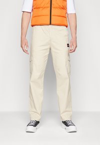 Pantalon cargo beige avec poches latérales, associé à un gilet matelassé orange. Le pantalon comporte un patch logo et est porté avec des baskets noires.