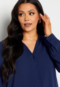 Camicia button-up blu navy con scollo a V, maniche lunghe e tessuto testurizzato. Include bottoni e polsini delle maniche leggermente a sbuffo.
