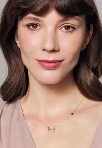 Złoty naszyjnik z okrągłym zawieszeniem i napisem, z delikatnym łańcuszkiem. Tło w neutralnym odcieniu, podkreślające projekt biżuterii.