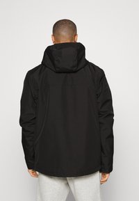 Chaqueta impermeable negra con capucha, que presenta una textura suave y un corte holgado. La parte trasera muestra un diseño simple sin logotipos visibles.