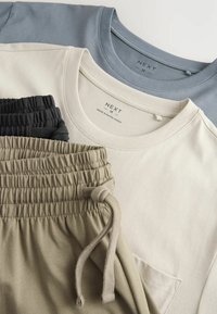 Tre T-shirts i blått, beige och krämfärgat staplade ovanpå ljus olivgröna och svarta joggers med elastiska midjeband och snörning. Mjuka tygtexturer.