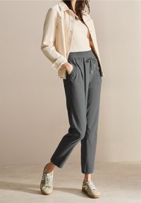 Pantalon de jogging gris avec taille élastique, poches latérales et coupe décontractée, associé à une veste beige clair et des baskets décontractées.