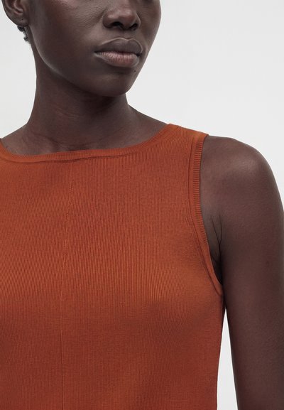 Κοντινό πλάνο ενός ατόμου με σκούρο δέρμα που φοράει αμάνικη, ριγωτή, σκουροκόκκινη μπλούζα σε ανοιχτό γκρι φόντο.