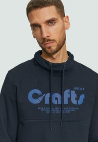 Navyblauwe pullover met een hoge kraag, voorzien van een groot bedrukt "Crafts" logo in lichtblauw. Met trekkoorden en een gladde textuur.