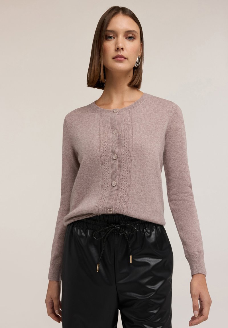 Cardigan roz deschis cu butoni, având un panou frontal texturat și mâneci lungi; asortat cu pantaloni jogger negri, lucioși, cu talie elastică.