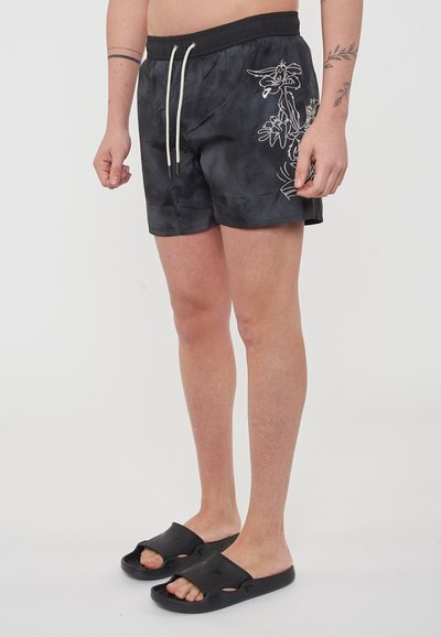 Pantaloni da bagno neri con effetto tie-dye, dotati di cintura elastica e cordoncino, con una stampa bianca di fiori e personaggi, abbinati a sandali neri.