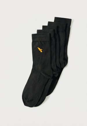 Penki juodų kojinių su raštu, suslankiusių į krūvą, kiekvienos kojinės viršuje šalia apvado matomas mažas geltonas išsiuvinėtas paukštelis, baltame fone.