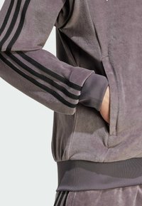Persona vistiendo un chándal de terciopelo púrpura con rayas negras, metiendo la mano en el bolsillo delantero, primer plano del torso y el brazo.