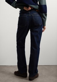 Jeans de mezclilla azul oscuro con piernas rectas, bolsillos traseros visibles y una etiqueta de cuero en la cintura. Llevados con zapatos marrones.