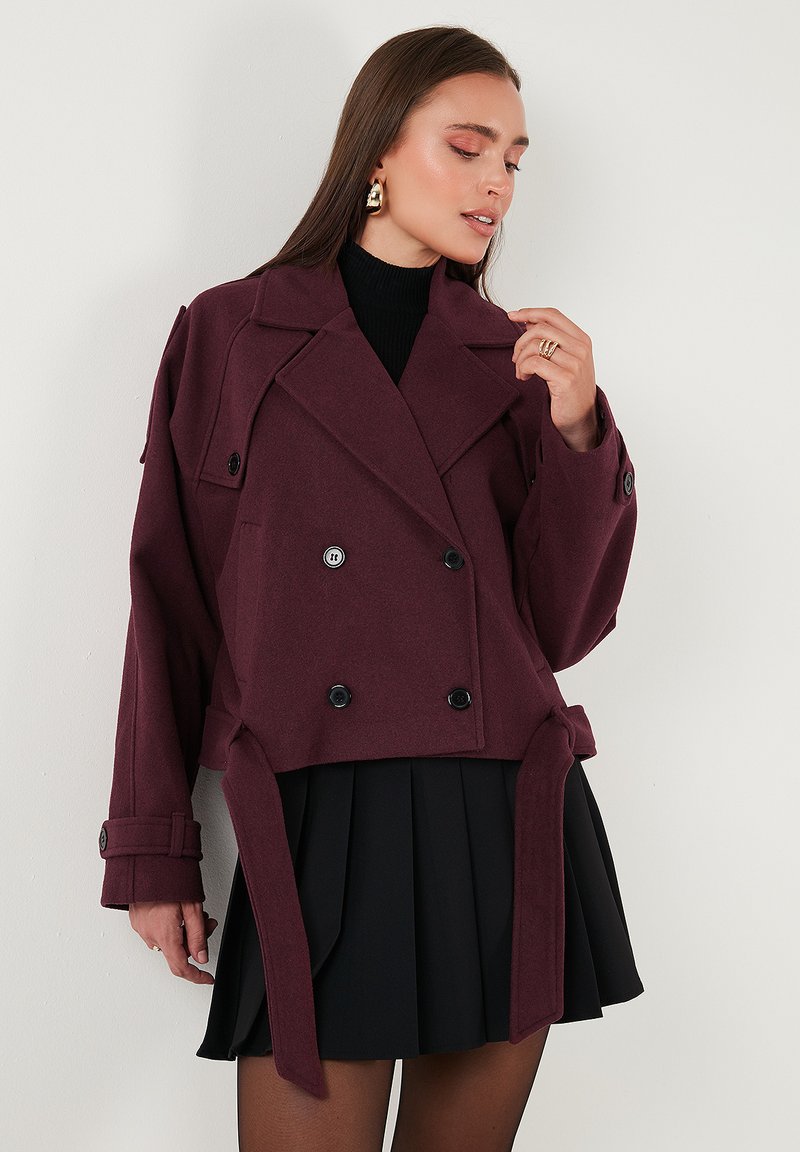 Burgundská dvojřadá cropped bunda s velkými klopami, ozdobnými knoflíky a zavázaným pasem. Nositá přes černou plisovanou sukni.
