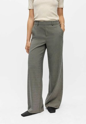 Femme portant un haut beige à manches courtes et un pantalon gris taille haute à jambes larges, chaussures noires, mains dans les poches, sur fond blanc.