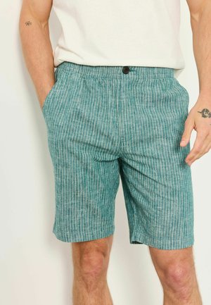 Mann trägt türkisgrüne Leinen-Shorts mit dünnen weißen vertikalen Streifen und ein weißes, hineingestecktes Hemd, Hände in den Taschen.