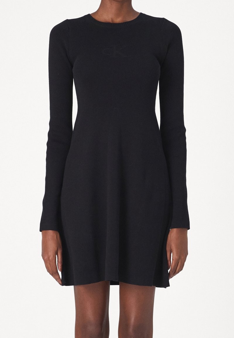 Femme portant une robe noire cintrée à manches longues avec une texture côtelée subtile et un léger évasement à l’ourlet, debout devant un fond uni.