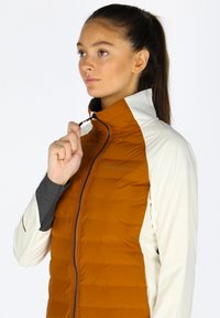 Isoleret jakke med en orange-brun puffy krop, cremede ærmer, høj krave og lynlåsdetalje. Minimalistisk design, kontrasterende farver, let tekstur.