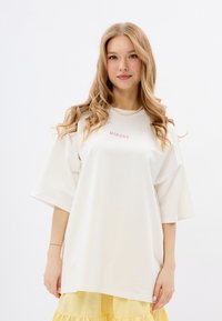 Oversized witte t-shirt met korte mouwen, voorzien van roze geborduurde tekst "MIRONS" op de borst. Gladde katoenen stof.