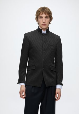 Ung mand med krøllet hår iført en sort blazer med mandarin-krave og mønster over en hvid skjorte og mørke bukser, stående foran en ensfarvet baggrund.