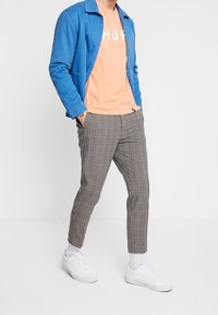 Blå jacka, orange T-shirt med vit text, grå rutiga byxor och vita sneakers. Mjuka texturer, modern passform och rena linjer.