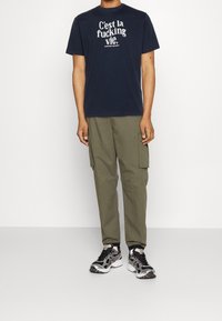 T-shirt en coton bleu marine avec texte blanc, pantalon cargo olive avec poches latérales et baskets multicolores. Tenue décontractée avec une coupe ample.