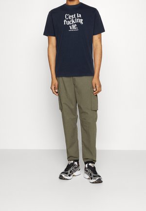 T-shirt en coton bleu marine avec texte blanc, pantalon cargo olive avec poches latérales et baskets multicolores. Tenue décontractée avec une coupe ample.