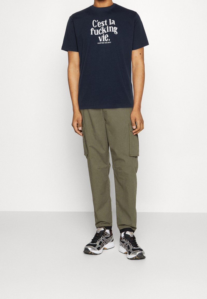 T-shirt en coton bleu marine avec texte blanc, pantalon cargo olive avec poches latérales et baskets multicolores. Tenue décontractée avec une coupe ample.