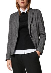 Blazer gris en chevrons avec une coupe ajustée, des revers crantés et un léger éclat. Porté sur un haut noir texturé et une chemise blanche.