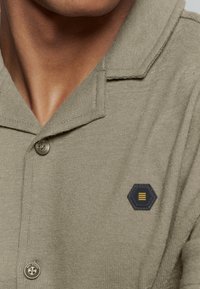 Khaki kortgebouwde gebreide shirt met een textuurfinish, kenmerkend door een zwarte hexagonale patch met gele accenten en een knoopsluiting bij de kraag.