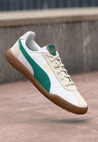 Valge ja roheline Puma toss, mis hõljub tekstureeritud pinna kohal, näidates oma disainidetaile, sealhulgas brändingut ja paelu.