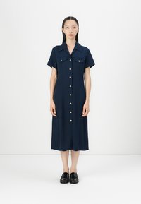 Marineblå knapperopkjole med korte ærmer, der har to brystlommer og guldtonede knapper, lavet af et glat stof. Båret med sorte loafers.