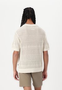 Maglione corto a maniche corte di colore crema lavorato a maglia con motivo a rete aperta e colletto a coste, indossato con pantaloni corta khaki. Vista posteriore che mette in mostra la texture e il design.