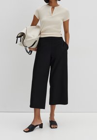 Kremowa bluzka z krótkim rękawem i dekoltem w kształcie łezki, w połączeniu z luźnymi czarnymi kulotami i czarnymi sandałami na blokowym obcasie, trzymająca beżową torbę.