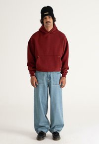 Mand iført en sort beanie, maroon hoodie, løse lyseblå jeans med store frontlommer og sorte sko, stående mod en ensfarvet baggrund.