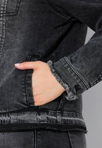 Mustaa farkkutakkia, jossa on risaset hihansuut ja helma, ja käsi sivutaskussa. Kankaassa on teksturoitu, hieman kulunut ilme.