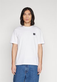 Tricou alb din bumbac, cu mâneci scurte, guler rotund și un mic patch cu logo negru pe partea stângă a pieptului. Purtați cu pantaloni din denim albastri.