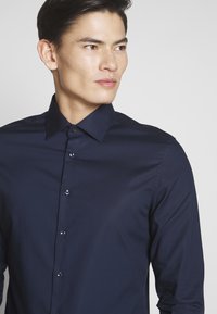 Mann mit kurzen dunklen Haaren, der ein eng anliegendes dunkelblaues Hemd mit Knöpfen trägt und leicht nach rechts blickt, vor einem schlichten hellen Hintergrund.