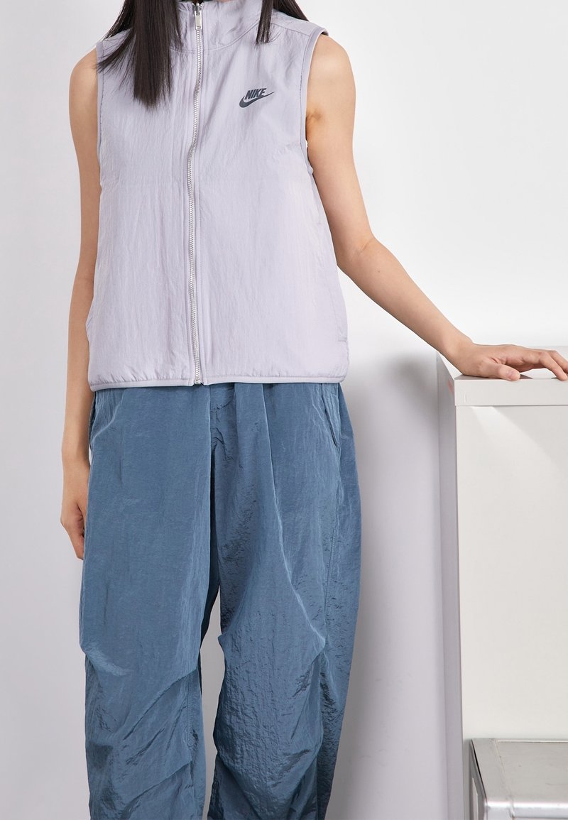 Ljuslila Nike ärmlös väst med dragkedja. Mjuk, strukturerad tyg. Tillsammans med lösa, blå byxor med en veckad design.