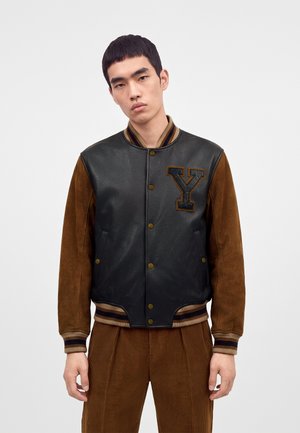 Jeune homme portant une veste varsity en cuir noir avec des manches en suède marron et une grande lettre Y sur la poitrine, associée à un pantalon en velours côtelé marron.
