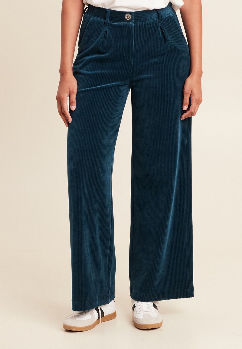 Pantalons en velours côtelé à jambes larges de couleur bleu profond, avec des côtes verticales visibles, une fermeture à bouton à l'avant et des plis. Associés à des baskets blanches.