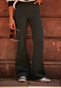 Pantalones de campana en color carbón, confeccionados con tela suave, que presentan una cinturilla lisa y costuras detalladas. Se combinan con zapatillas plateadas y sosteniendo unas gafas de sol.